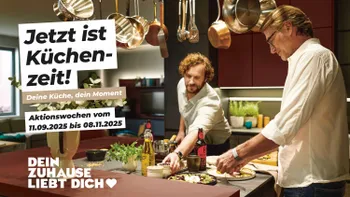 Zwei Männer kochen entspannt gemeinsam in moderner Küche mit roten Fronten.